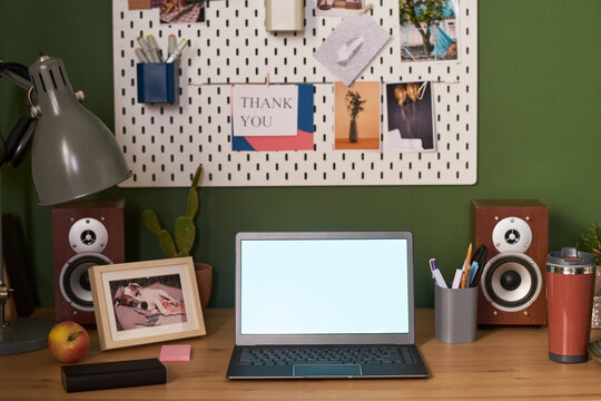 Modern workspace featuring open laptop with blank screen on wooden desk, framed photo, speakers, cactus, apple, reusable cup, organized pegboard with thank you card and photos in background