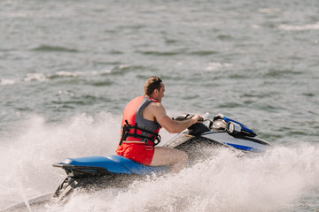 Obraz premium Male rider in red swim shorts operates a blue jet ski on a lake, splashing water as he navigates through the waves under a clear sky