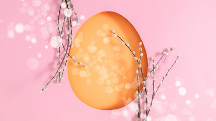 A close-up of an Easter composition featuring a silhouette of a large orange paper egg on a soft pink background with bokeh highlights, complemented by willow branches in a flat lay.