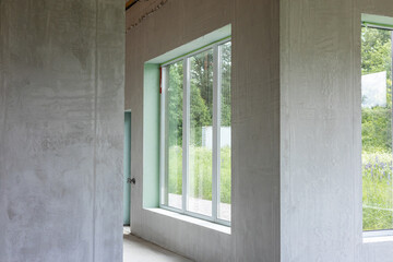 Obraz premium Interior of an unfinished room with large modern windows. Bare concrete walls and floor, natural light, and green trees view. A construction or renovation project.