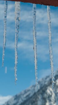 Motion Controlled Vertical Time Lapse of melting icicles hanging from roof with Snowy Mountain Peak and Moving Clouds in the background. Tripod shot