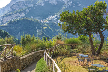 Amalfi Coast, Italy. Scenic view of a winding pathway leading to outdoor seating area with metal tables and chairs surrounded by lush greenery and mountains in the background