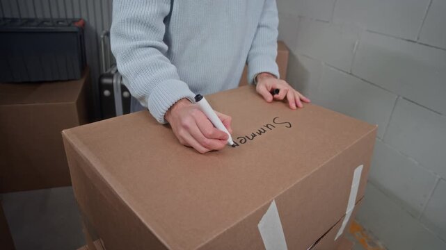 Middle aged man writing Summer on cardboard box, then lifting it and handing to his son while organizing personal belongings inside secured self storage unit