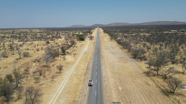 Drone aerial view in Namibia desert flying over a narrow road following a SUV car with sand and green trees on the sides and mountains in the horizon on a sunny day