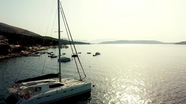 Sailboat yacht catamaran anchored near shoreline with sun reflections shimmering across calm sea surface, aerial dolly