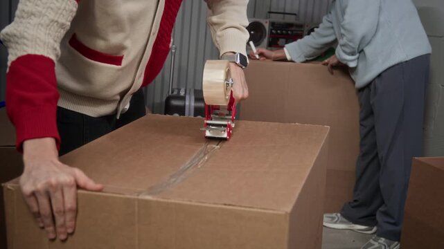 Close up shot of unrecognizable man sealing cardboard box with packing tape dispenser while another man writing label on different box in storage room during moving process