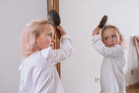 Cute little girl brushing her hair in front of the mirror at home.