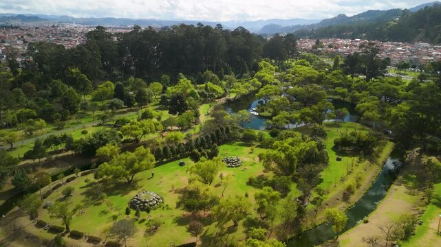 Reveal a wide view of the peaceful park lake, lush green trees, and walking paths in Parque El Paraiso under bright daylight in Azuay - aerial rising while tilting down