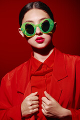 Fashionable woman in vibrant green round sunglasses and bold red lips poses in a red outfit on red...