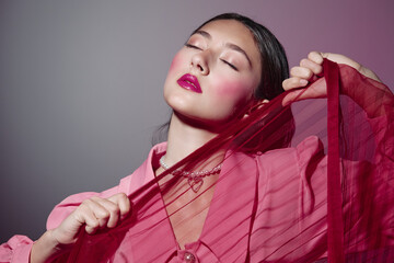 Young woman with glossy pink lips and vibrant blush poses holding a red sheer fabric against grey...