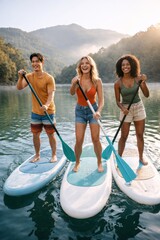 Obraz premium Group of young friends paddle boarding together on beautiful lake surrounded by mountains and sunshine