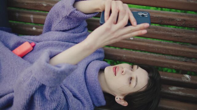 Young woman lies on bench and looks at phone in park during sunny day while wearing purple sweater and holding pink object