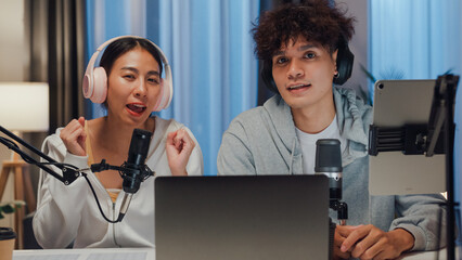 Young happy Asian couple wear headphone sit on desk online live streaming football live score looking at camera watch soccer sport game on laptop talk with follower in small studio at home at night.