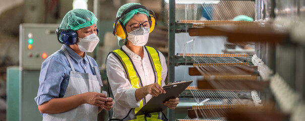 Textile inspection factory teamwork female supervisor in safety vest checking and discussing yarn production quality with woman worker in textile mill holding thread spool and clipboard. © BESTIMAGE