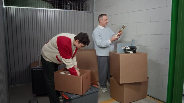 Rack focus shot of middle aged man and his young son unpacking cardboard boxes inside storage unit, examining golden trophy and reminiscing together