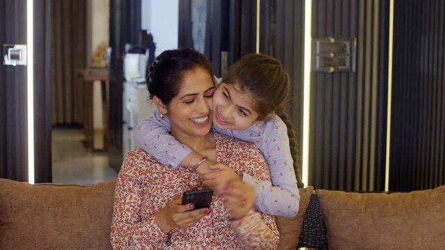 An attractive Indian housewife in casual clothes browsing / scrolling on mobile - busy mother  single mother  modern housewife. A little girl surprises her mother by giving her a hug while she is s...