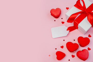 Gift box tied with red ribbon, heart-shaped macarons, and a blank tag arranged on a pink background, symbolizing love, romance, and celebration for special occasions