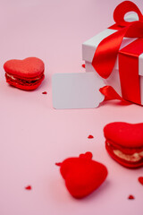 White gift box with red ribbon and blank tag presenting heart-shaped macarons and scattered confetti on a romantic pink background, symbolizing love and celebration for Valentine's Day