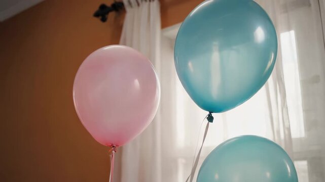 A hand holds a pink balloon next to a blue balloon. The background features soft curtains and a warm-colored wall, symbolizing a baby shower or gender reveal celebration.