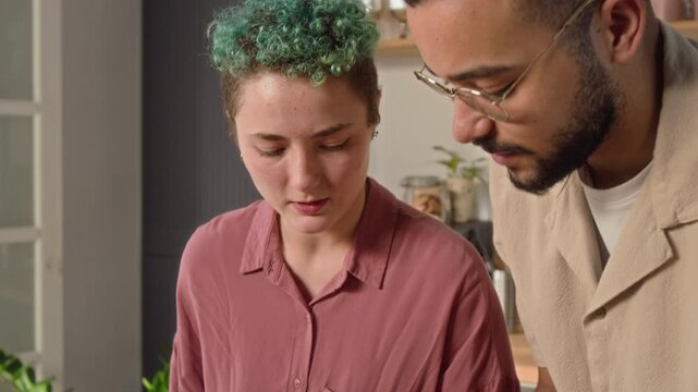 Tilt up shot of positive young couple viewing pizza recipe on digital tablet and discussing it, man showing how to cut tomato with gesture and playfully throwing it up