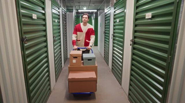 Tilt up full length shot of young man pushing cart filled with various cardboard boxes while walking through hallway of storage facility and examining green metal doors of units