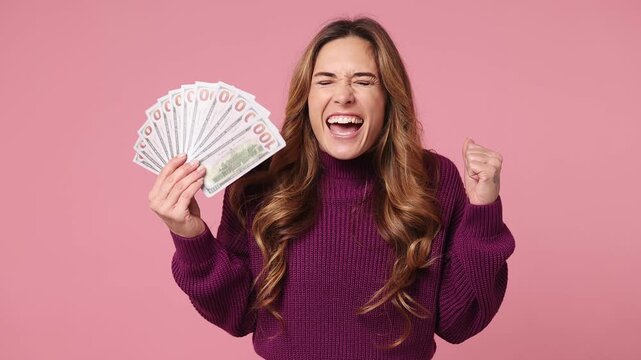 Happy fun young woman she wear purple knitted sweater casual clothes hold in hand fan of cash money in dollar banknotes doing winner gesture celebrate isolated on plain pastel light pink background