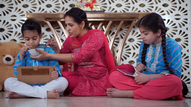An Indian mother feeding noodles to her son - using a tablet  screen addiction  tasty meal  online learning. Cute Indian siblings are using a digital tablet while sitting with their mother - a Punj...