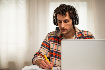 Focused young man wearing wireless headphones, working on his laptop at home while taking notes on a notebook.	
