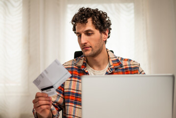 Modern young man using credit card and laptop at home. He is focusing on e-commerce and managing his finances.	