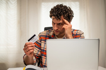 Young businessman using laptop for online banking, holding his credit card and looking worried...