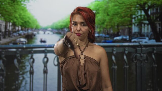 Woman in brown dress shows thumbs down with extended hand on a street bridge over a canal in amsterdam; defiance protest.