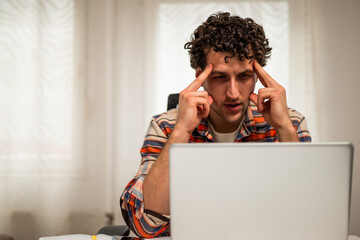 Despaired and exhausted young businessman having a headache while working on laptop at his home.	