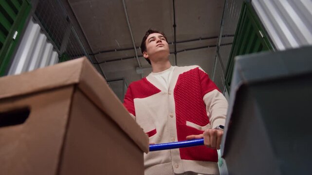 Low angle view of young man walking and pushing cart filled with cardboard boxes through hallway in storage facility, looking at metal doors while searching for his unit
