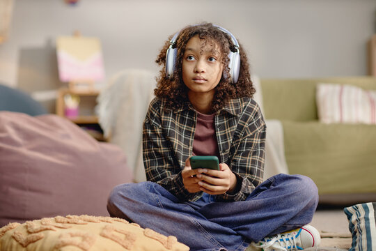 Portrait of Black teenager girl sitting cross legged holding smartphone wearing large headphones looking upward indoors representing Generation Alpha using modern gadget