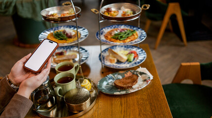 Fototapeta premium Person holding a smartphone while enjoying a tiered tray of food and tea