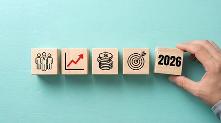 Hand placing 2026 block with team growth money and goal icons on light blue for business planning and future success