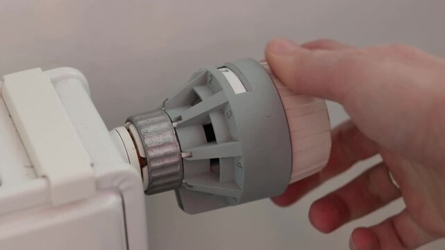 A woman's hand turns a mechanical thermostat on a radiator. A close-up of a temperature regulator on a battery. A mechanical thermal head of a battery regulator with a scale from 0 to 5
