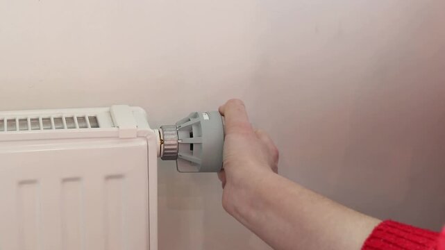 A woman's hand turns a mechanical thermostat on a radiator. A close-up of a temperature regulator on a battery. A mechanical thermal head of a battery regulator with a scale from 0 to 5