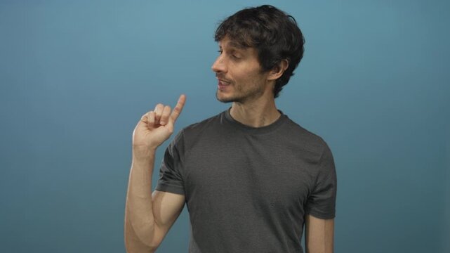 Young man with raised index finger pointing gesture and closed eyes in blue studio with slight smile; contemplation.