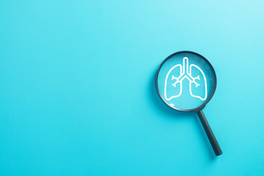 Magnifying glass focusing on white human lungs icon on blue background with copy space. Respiratory health checkup, lung cancer diagnosis, pneumonia virus, and air pollution protection concept.