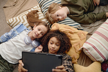Three children lying on pillows using digital tablet, multiethnic group including Caucasian boy,...