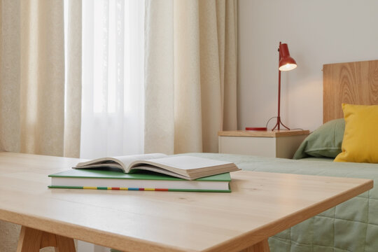 Stack of books with open notebook lying on wooden desk in front of bed with green bedding and yellow pillow, sunlight streaming through window with curtains in background