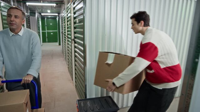 Middle aged man pushing cart down corridor of self storage facility while another tenant unloading cardboard boxes from his unit