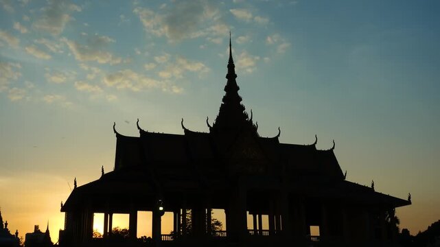Timelapse silhouette of ornate royal palace temple roof at sunset as clouds drift across the sky, cinematic travel landmark background with dramatic spire and warm twilight glow.