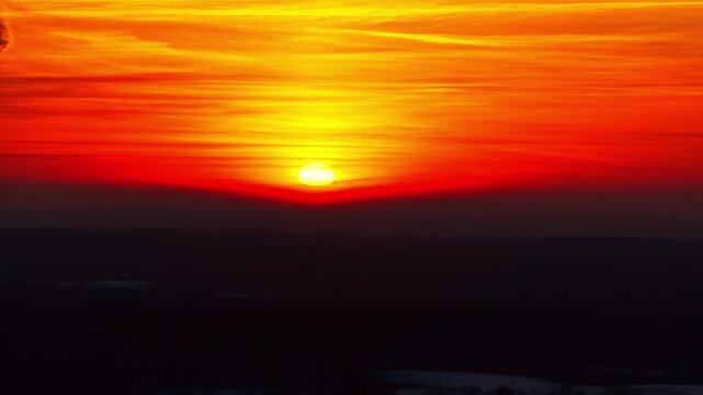 Fiery winter sunrise close-up with sun touching the horizon, intense orange clouds and dark silhouette foreground, dramatic sky background for cinematic and abstract use.