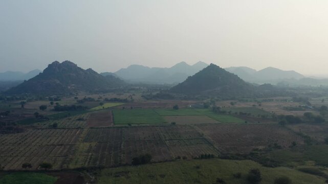 Wide aerial view of cultivated farmland and layered rocky hills under hazy light near Warangal, Telangana, India, showing rural scenery, agriculture patterns, and natural terrain.