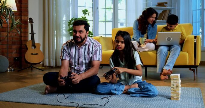 Indian father daughter playing video game and mother son sitting on sofa behind as family enjoys fun time together indoors in modern lavish home living room lifestyle scene
