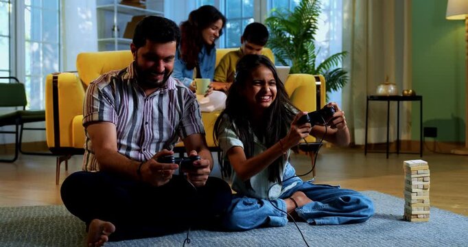 Indian father daughter playing video game and mother son sitting on sofa behind as family enjoys fun time together indoors in modern lavish home living room lifestyle scene