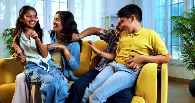 Indian young parents tickling kids at home while sitting on sofa in playful living room, happy father and mother laughing with son and daughter indoors, joyful family lifestyle moment