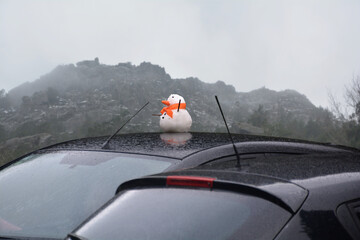 Little snowman on top of a parked car roof, with snowy mountains in the background, winter and cold